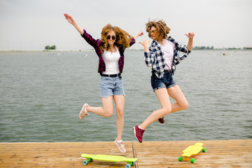 Two cheerful happy skater girls in hipster outfit having fun on a wooden pier during summer vacation
