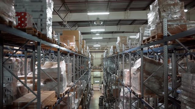 A large warehouse with boxes of goods. The camera moves up along the shelves with cardboard boxes. A forklift works in the distance