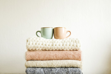 Fototapeta na wymiar Two cups of coffee standing on bunch of knitted warm pastel color sweaters w/ different knitting patterns folded in stack. Fall winter knitwear clothing. Textured wall background. Close up, copy space