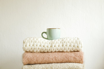 Cup of cappuccino coffee on bunch of knitted warm pastel color sweaters w/ different knitting patterns folded in stack. Fall winter knitwear clothing. Textured wall background. Close up, copy space.