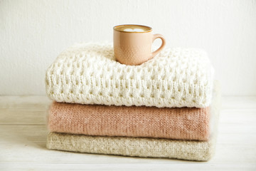 Fototapeta na wymiar Cup of cappuccino coffee on bunch of knitted warm pastel color sweaters w/ different knitting patterns folded in stack. Fall winter knitwear clothing. Textured wall background. Close up, copy space.
