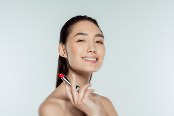 beautiful smiling asian woman holding red lipstick, isolated on grey