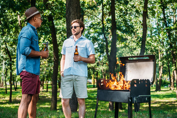 multiethnic friends with beer having conversation near grill in park