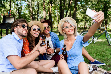 smiling multicultural friends with beer taking selfie on smartphone while resting in summer park