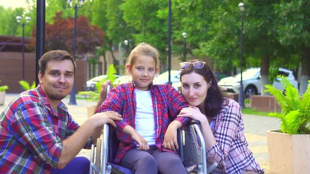 portrait of looking at the camera mom and dad with a little daughter disabled in a wheelchair, happy
