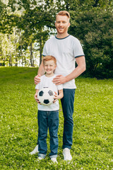 happy father and son with soccer ball standing together and smiling at camera in park