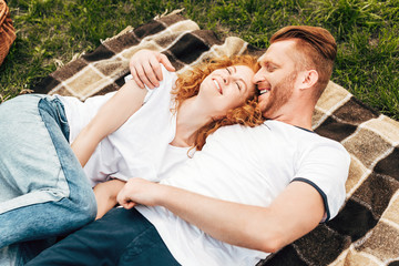 high angle view of happy young redhead couple hugging and lying on plaid at picnic