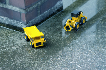 There are excavator and truck toys next to waterlogged cement and old brick pillars.