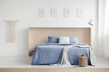 Blanket on blue bed with pillows in white bedroom interior with posters and lamp. Real photo