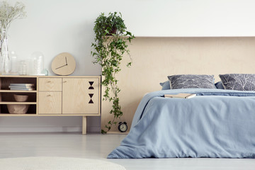 Clock on wooden cupboard next to to blue bed with patterned pillows in bedroom interior. Real photo