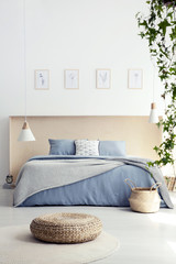 Pouf in front of blue bed with blanket in minimal bedroom interior with lamp and posters. Real photo
