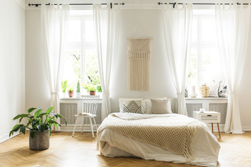 Plant and white chair next to bed with blanket in bright bedroom interior with windows. Real photo
