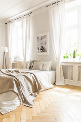 Beige blanket on bed and poster in white bedroom interior with drapes at windows. Real photo