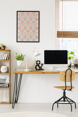 Poster on white wall above wooden desk with lamp and desktop computer in workspace interior. Real photo