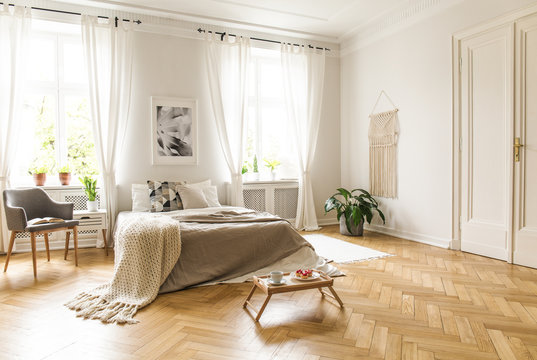 Grey armchair next to bed with blanket in bright bedroom interior with poster and windows. Real photo