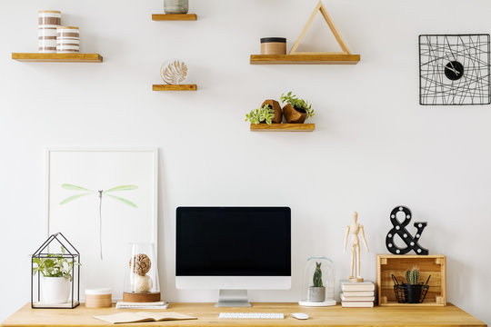 Poster and computer desktop on wooden desk in white home office interior with shelves. Real photo