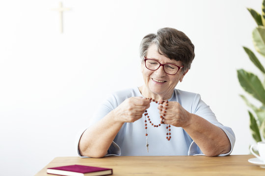 Smiling senior woman holding rosary while sitting at table with bible - Powered by Adobe