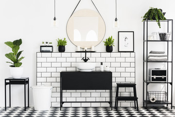 Mirror between plants above black washbasin in bathroom interior with checkered floor. Real photo