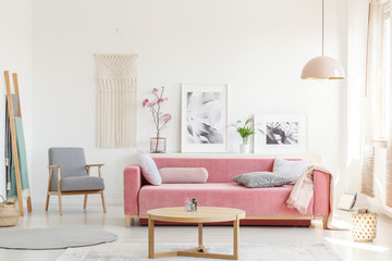Patterned armchair next to pink sofa in apartment interior with posters and wooden table. Real photo