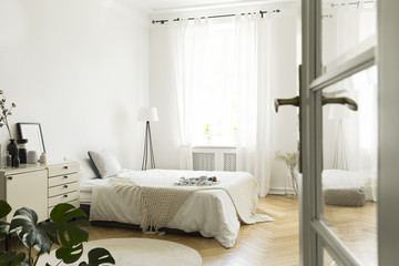 Knit blanket on bed in white bedroom interior with lamp next to window. Real photo