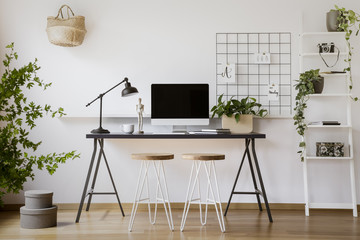 Stools at desk with lamp and computer desktop in freelancer's interior with plants. Real photo