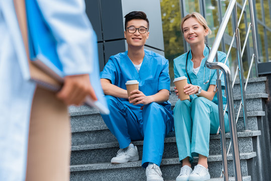 cropped image of teacher looking at smiling medical students sitting on stairs with coffee to go