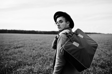 Stylish man in glasses, brown jacket and hat with bag posed on green field.