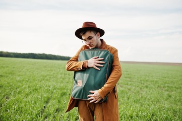 Obraz premium Stylish man in glasses, brown jacket and hat with bag posed on green field.