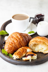 Breakfast concept: two fresh french croissant with a cup of coffee and jam on a dark wooden tray.