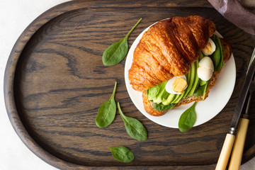 Croissant with green salad, avocado and eggs on a dark wooden tray. Breakfast, brunch or lunch concept.