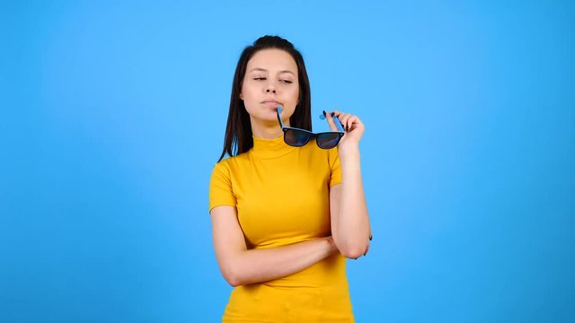 Positive, happy and bored young woman plays with her sunglasses on blue background. Fashionable teeanger. Slow motion footage