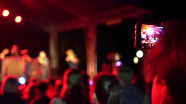 Female fan at concert holds smartphone in hand and takes picture or shoots video in multi colored background blurred performance of singer and musical group