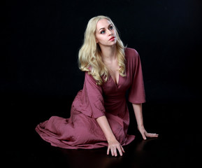 full length portrait of blonde girl wearing long purple dress. seated pose. black studio background.