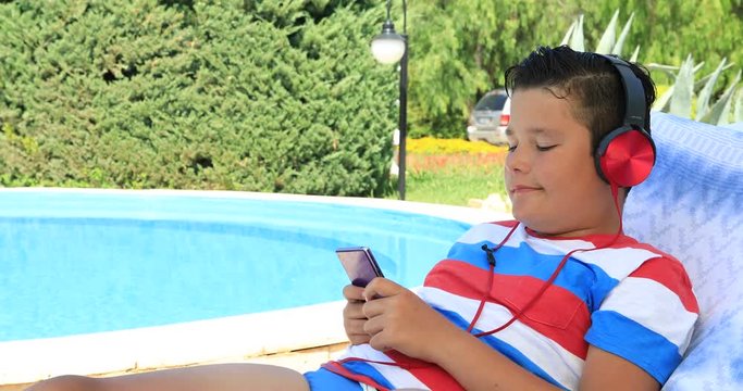 Portrait of a cute teenage boy with headphone sitting on a sunbed near the swimming pool and using smartphone on a sunny summer day.