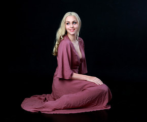 full length portrait of blonde girl wearing long purple dress. seated pose. black studio background.
