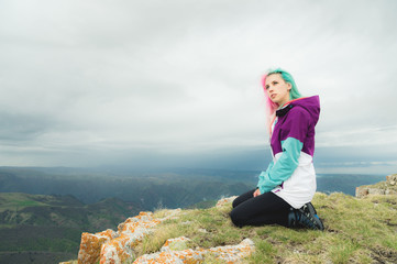 A girl-traveler with multicolored hair sits on the edge of a cliff and looks to the horizon on a background of a rocky plateau