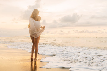 Hipster trendy woman in casual summer dress walk barefoot by the waterline and look to the little waves. Sporty lady on sea sand beach sunset or ocean sunrise. Travel, active, yoga lifestyle concept. - 214243550