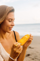 Healthy vegetarian hipster woman in summer casual outfit with fresh grilled corn. Sexy lady on sea beach sunset or ocean sunrise. Travel, explore, active yoga and meditation lifestyle concept.