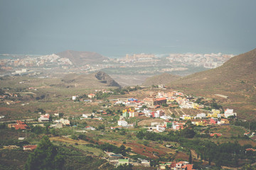 Fototapeta na wymiar An elevated view over a Tenerife village of brightly coloured houses, the surrounding landscape and hills, the coast and ocean, all covered with the haze of La Calima (coastline of Costa Adeje)