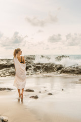 Pretty girl stay in front of huge waves crashing on the rocks. Young lady on exotic beach seaside enjoying sunset or ocean sunrise. Danger, warning, travel, surfer relax and active lifestyle concept.