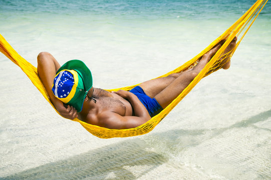 Brazilian man in sunga bathing suit and brazil flag hat relaxing in seaside beach hammock in Brazil. - Powered by Adobe