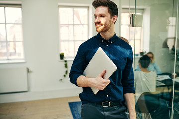 Smiling young businessman walking with a laptop in an office - obrazy, fototapety, plakaty