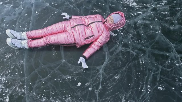 Girl flies and rolls on the ice. Young couple has fun during winter walk against background of ice of frozen lake. Lovers lie on clear ice with cracks, have fun, kiss and hug. View from above. Young
