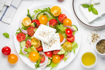 Greek salad with colorful cherry tomatoes red and yellow, cucumber, onion, lettuce and large piece of feta cheese with herbs. In white plate on light background