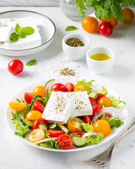 Greek salad with colorful cherry tomatoes red and yellow, cucumber, onion, lettuce and large piece of feta cheese with herbs. In white plate on light background