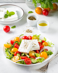 Greek salad with colorful cherry tomatoes red and yellow, cucumber, onion, lettuce and large piece of feta cheese with herbs. In white plate on light background