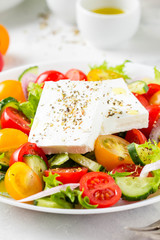 Greek salad with colorful cherry tomatoes red and yellow, cucumber, onion, lettuce and large piece of feta cheese with herbs. In white plate on light background