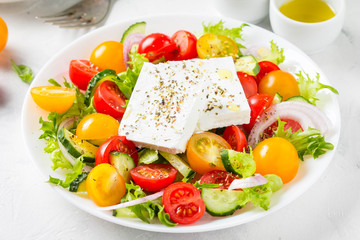 Greek salad with colorful cherry tomatoes red and yellow, cucumber, onion, lettuce and large piece of feta cheese with herbs. In white plate on light background