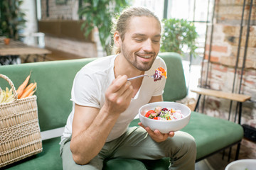 Handsome man eating healthy salad sitting indoors on the green sofa with bag full of vegetables on...
