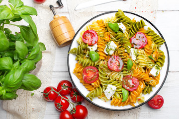 Włoskie jedzenie - Sałatka z kolorowym makaronem, pomidorami koktajlowymi, serem feta i świeża bazylią na białym drewnianym tle.   - obrazy, fototapety, plakaty